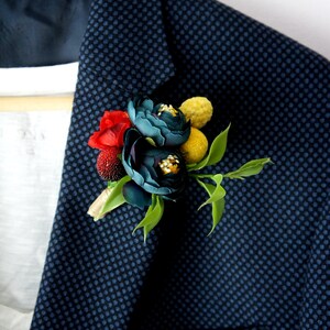 May include: A navy blue blazer with a small dot pattern, adorned with a floral boutonniere. The boutonniere features teal and red flowers, yellow accents, and green leaves. The blazer is worn over a white shirt.