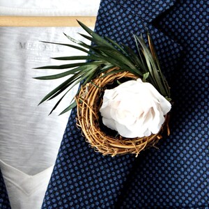 May include: A white rose with a woven twig base and green leaves, pinned to a blue patterned blazer.