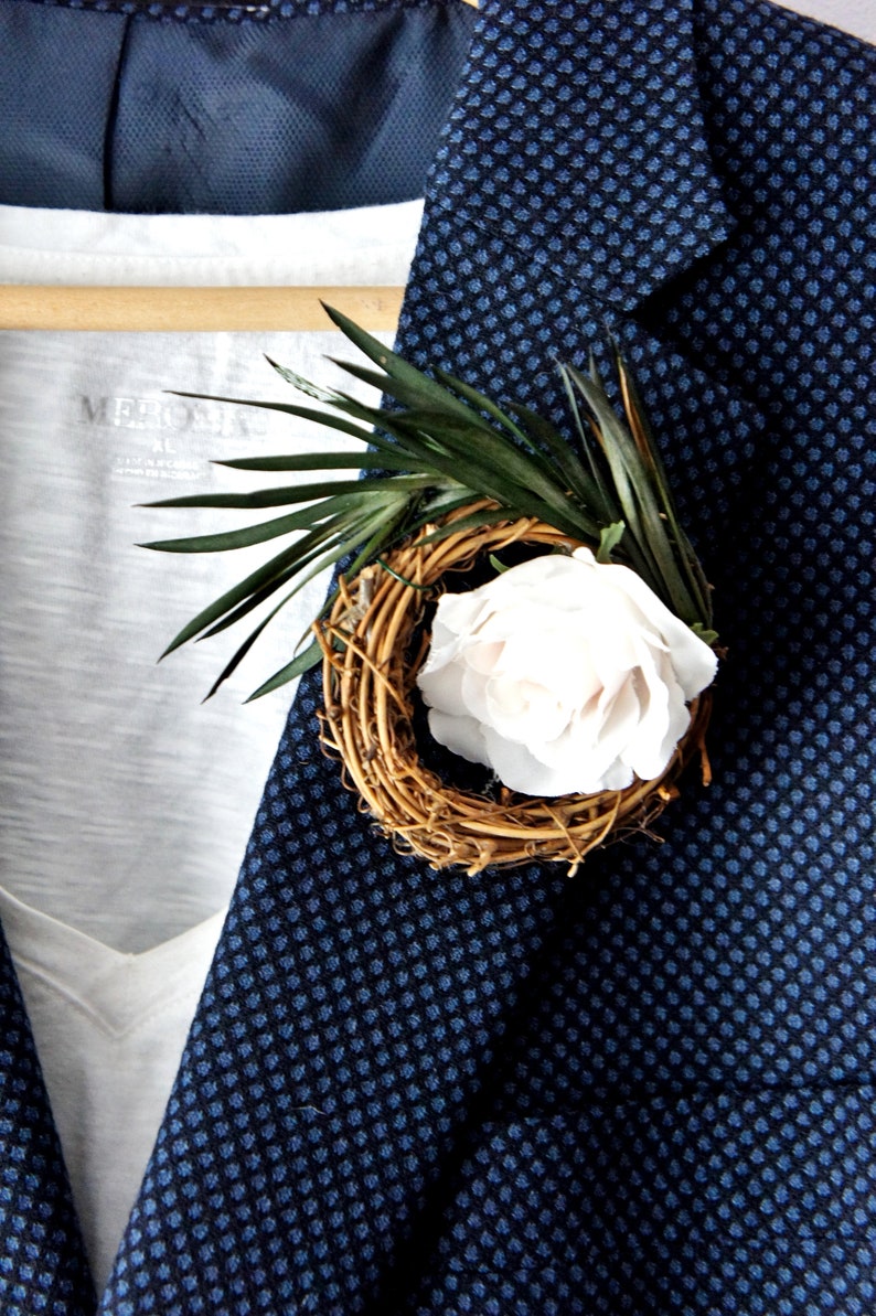 May include: A white rose with a woven twig base and green leaves, pinned to a blue patterned blazer.
