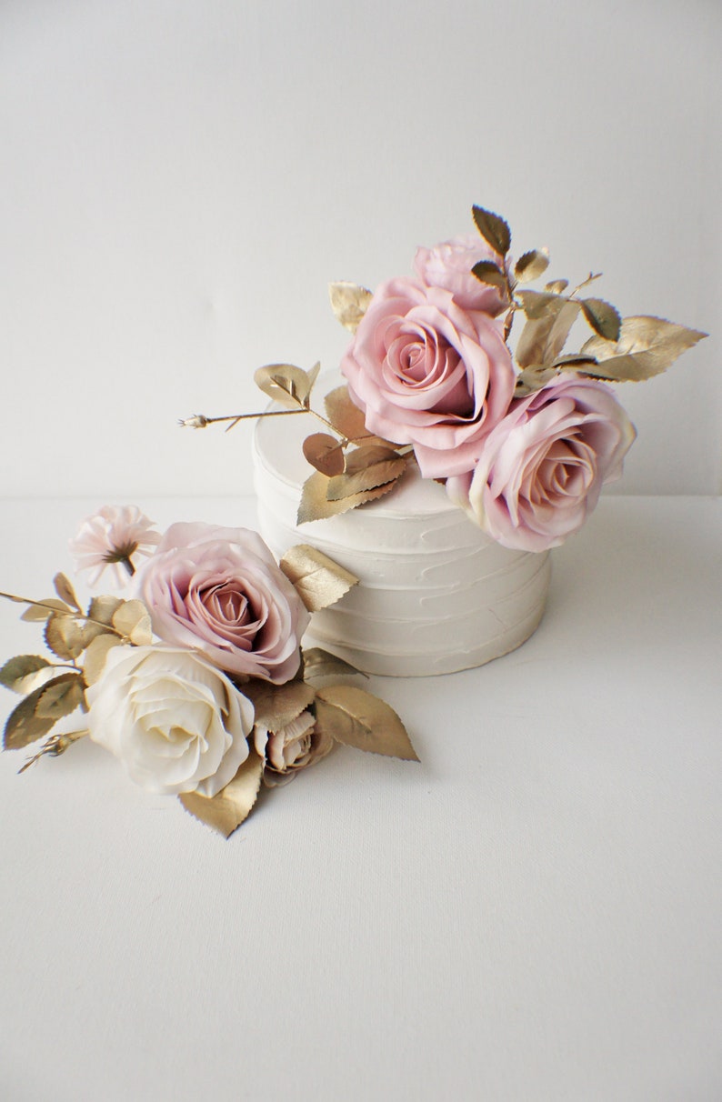 May include: Two arrangements of artificial flowers, one on top of a white cake and one on a white surface. Both arrangements feature pink and white roses with gold-painted leaves.