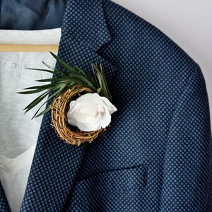 May include: A navy blue blazer with a small white flower and green leaves in a woven nest.