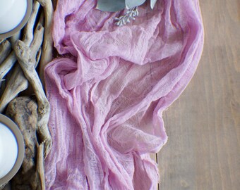 Könnte beinhalten: Ein rosa durchsichtiger Stoffläufer mit einer rustikalen Holzbasis und zwei weißen Kerzen. Der Stoff ist über das Holz gelegt und hat eine weiche, zarte Textur.