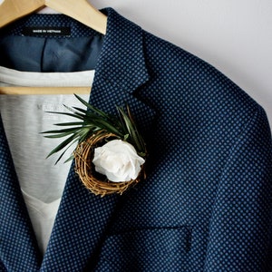 May include: A navy blue blazer with a small white flower and green leaves pinned to the lapel. The blazer has a subtle patterned design.