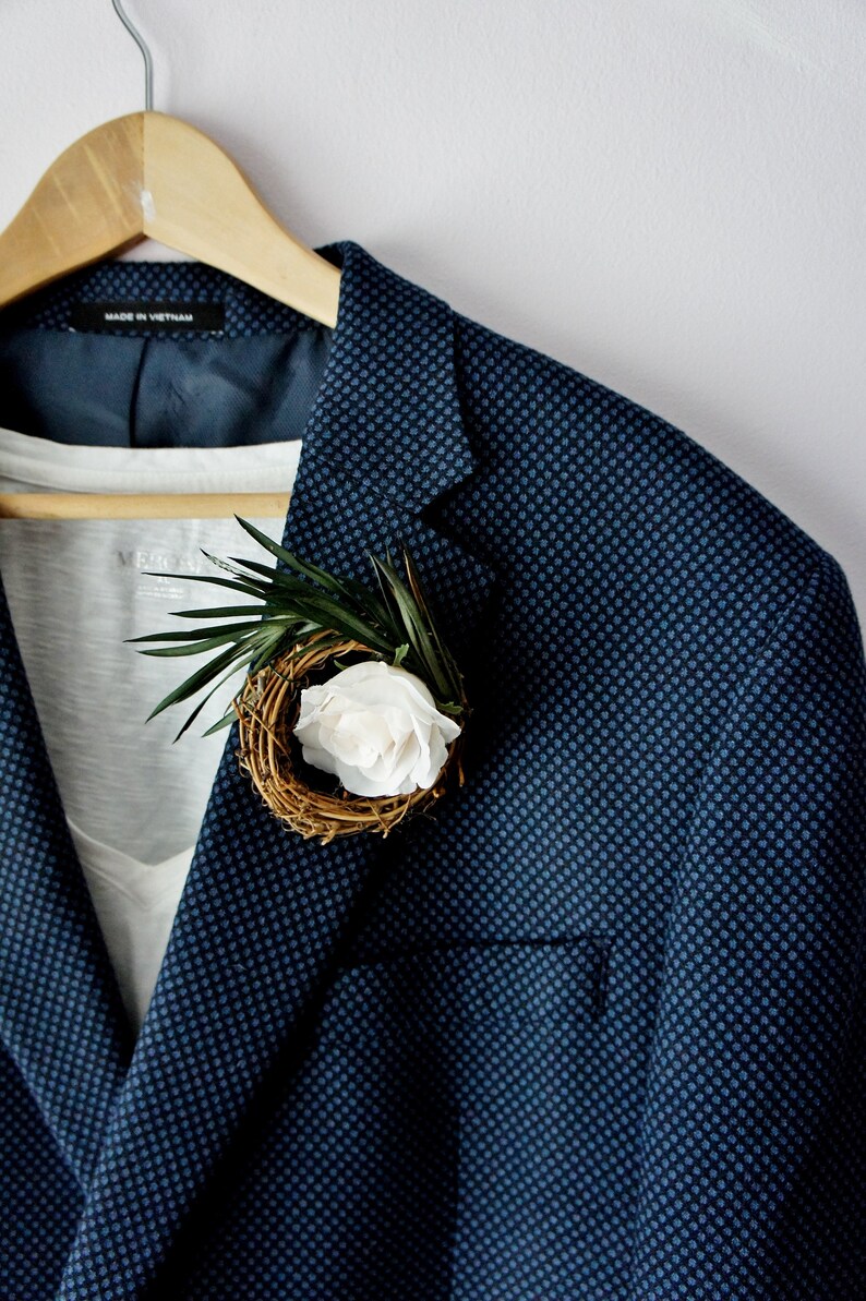 May include: A navy blue blazer with a small white flower and green leaves pinned to the lapel. The blazer has a subtle patterned design.