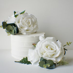 May include: White cake decorated with two white roses and greenery. The roses are made of fabric and the greenery is made of plastic.