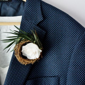 May include: A blue blazer with a small white flower and green leaves in a woven nest.