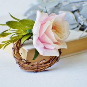 May include: A pink rose with green leaves is attached to a brown twig wreath.