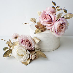 May include: Two white cake tiers decorated with pink and white roses and gold leaf accents. The cake is on a white surface.