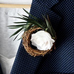 May include: A white rose in a woven twig wreath with green leaves, pinned to a dark blue patterned blazer.