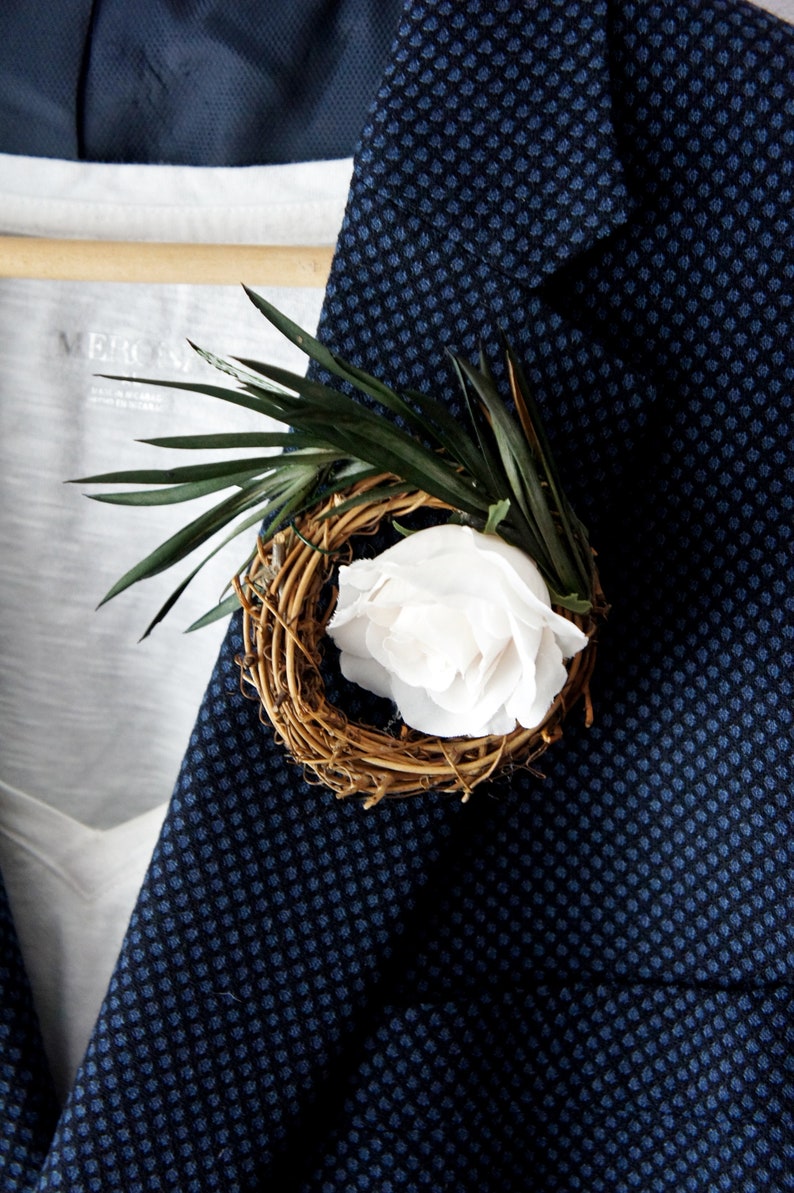 May include: A white rose in a woven twig wreath with green leaves, pinned to a dark blue patterned blazer.