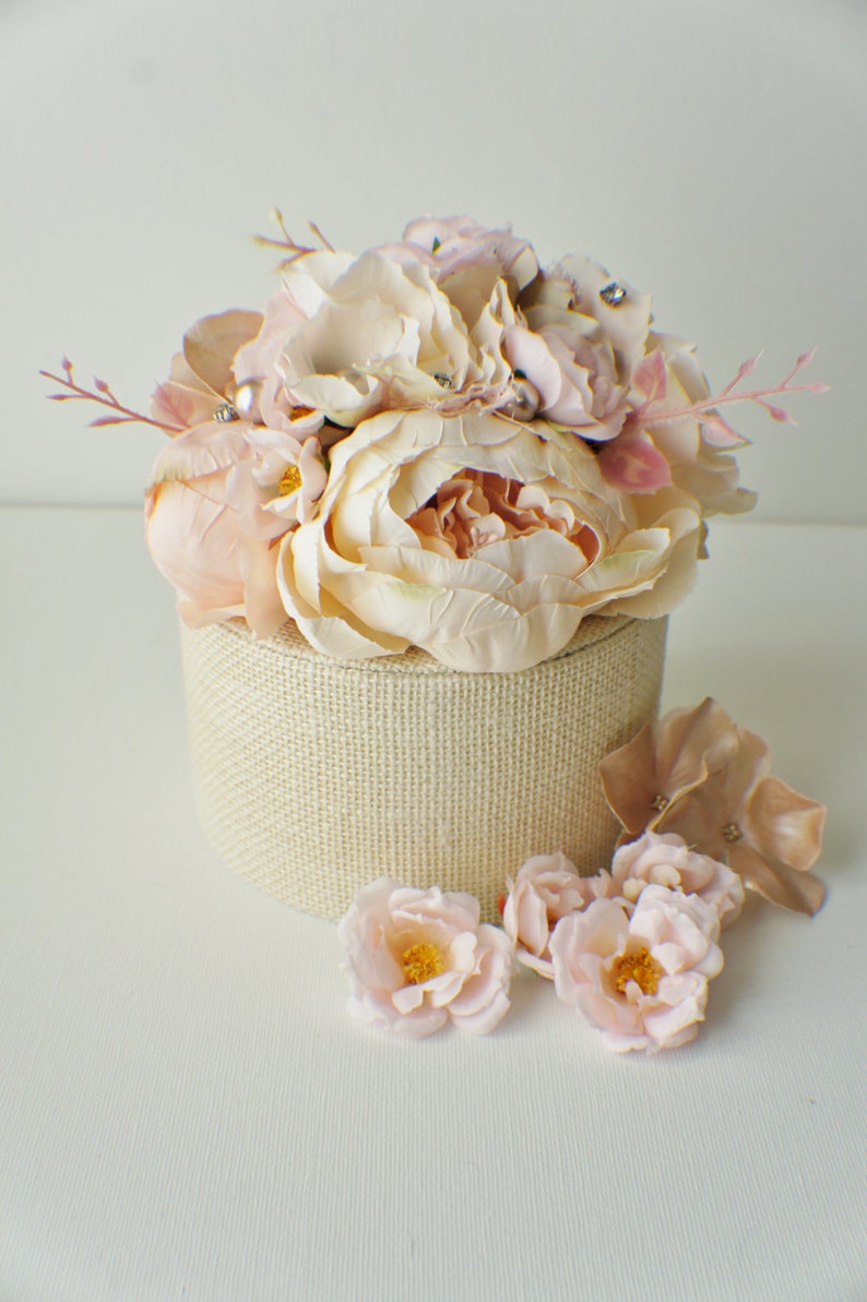 May include: A round, beige fabric-covered box topped with a cluster of light pink and cream-colored artificial peonies. Additional loose flowers and foliage are scattered around the base, creating a floral arrangement. The box is on a white surface.