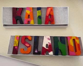 Puede incluir: Dos cajas que contienen cada una una palabra escrita con letras de crayones de colores. La caja superior deletrea "KAIA" con letras rojas, naranjas, amarillas, verdes y azules. La caja inferior deletrea "ISLAND" con letras rosas, naranjas, negras, blancas, verdes y amarillas.