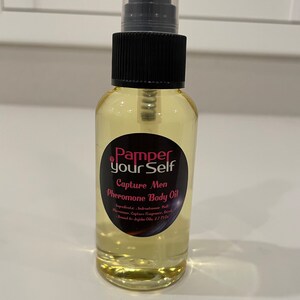 Może przedstawiać: Przezroczysta butelka szklana z czarną spryskiwaczką i różowo-czarną etykietą z napisem "Pamper Yourself Capture Men Pheromone Body Oil". Etykieta zawiera również listę składników i objętość 2,7 fl oz.