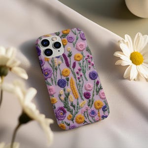 May include: A purple phone case with an embroidered floral design featuring yellow, pink, and purple flowers. The case has a camera lens cutout.