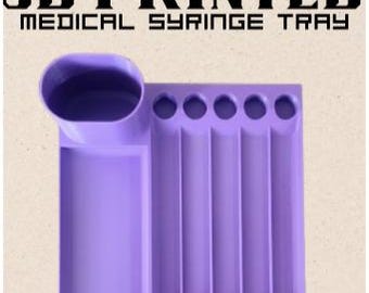 以下が含まれることがあります： ラベンダー色の3Dプリントされた医療用シリンジトレイ。円形のくぼみ、長方形の部分、5つの垂直スロットがあります。「3D PRINTED MEDICAL SYRINGE TRAY」の文字が黒で印刷されています。