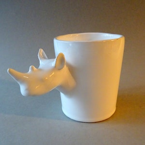 May include: White ceramic mug with a rhinoceros head handle. The mug is a simple cylindrical shape.