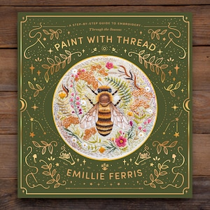 Puede incluir: Una cubierta de libro verde con letras doradas que dicen "A Step-by-Step Guide to Embroidery. Through the Seasons. Paint with Thread." La cubierta presenta una abeja bordada en un círculo de flores.