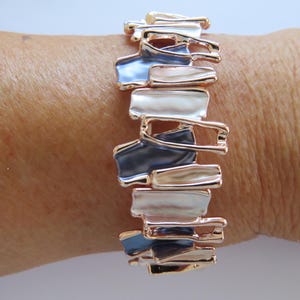 May include: A stretch bracelet featuring a series of interconnected, irregular rectangular metal links in rose gold tone. The links are accented with alternating enamel inserts in shades of glossy white, soft beige, and deep slate blue.