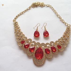 Op de afbeelding: Een gouden ketting en oorbellen set met rode en heldere strass steentjes. De ketting is een statement piece met een grote, druppelvormige hanger.