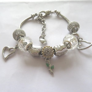 Puede incluir: Un brazalete de plata con una variedad de dijes, incluyendo corazones, un girasol y cuentas de vidrio transparentes.