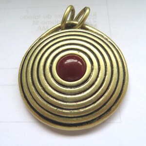 May include: Gold pendant with a red circular stone in the centre surrounded by concentric circles.