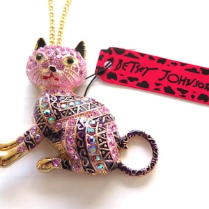 Può includere: Collana con pendente a forma di gatto rosa e oro con catena dorata. Il gatto è decorato con cristalli rosa e iridescenti e dettagli viola e neri. Un'etichetta rossa con "BETSEY JOHNSON" è attaccata.