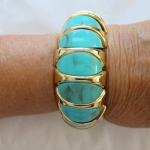 May include: A gold-toned bracelet with turquoise-colored, oval-shaped segments. The bracelet is worn on a wrist, and the segments are connected by gold-colored metal. The turquoise segments have a marbled appearance.