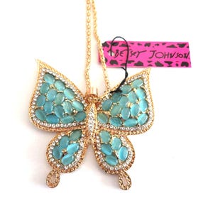 May include: Gold-toned butterfly pendant necklace with turquoise-colored stones and clear crystals. The butterfly design features a central row of crystals and turquoise stones in the wings. A pink tag with the text "BETSEY JOHNSON" is attached.