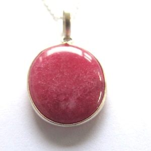 Könnte beinhalten: Runder, rosafarbener Edelsteinanhänger mit silberfarbener Metalleinfassung. Der Anhänger hängt an einer zarten Silberkette. Der Edelstein hat eine glatte, polierte Oberfläche und ein leicht gesprenkeltes Aussehen.