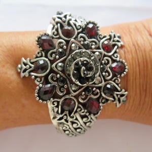 Peut inclure: Bracelet argenté orné d'un motif central en forme d'étoile avec un centre en spirale. Il est orné de pierres précieuses rouge foncé de forme ovale et de petites pierres noires. Le travail de filigrane complexe ajoute à son esthétique vintage.