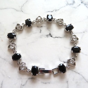 May include: A silver bracelet featuring black oval-shaped stones and clear heart-shaped stones. The bracelet has a silver clasp.