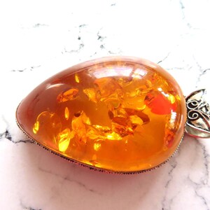 May include: A teardrop-shaped amber pendant with a silver bail. The amber is a deep orange colour and has inclusions of air bubbles and other organic matter. The pendant is approximately 5 cm long.