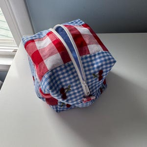 May include: A square, zippered pouch with a patchwork design. The pouch features red and white gingham fabric, blue and white gingham fabric, and cherry embroidery. The zipper is white, and the interior is blue.