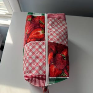 May include: A rectangular fabric pouch with a zipper closure. The pouch features a patchwork design with sections of red strawberries and red and white checkered patterns. A small strawberry tag hangs from the zipper.