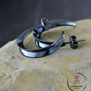 Könnte beinhalten: Ein Paar schwarze Creolen mit leicht gekrümmtem Design. Die Ohrringe sind aus Metall und haben eine polierte Oberfläche.