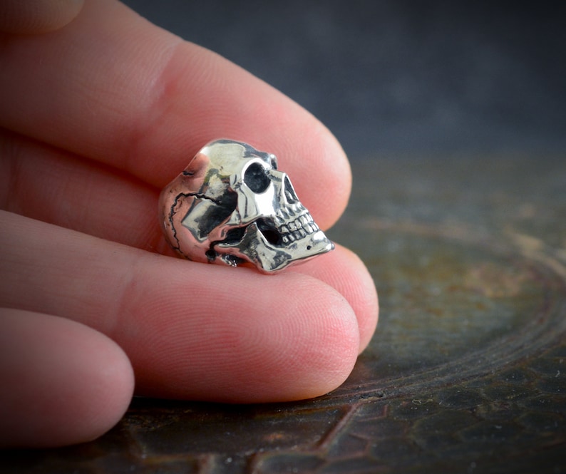 Op de afbeelding: Een zilveren schedelkraal met gedetailleerde kenmerken, waaronder tanden en oogkassen. De kraal wordt in close-up getoond, vastgehouden in een hand.