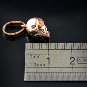 May include: A gold-colored skull pendant with green eyes, measuring approximately 1 cm in length. The pendant is shown next to a ruler for size reference.