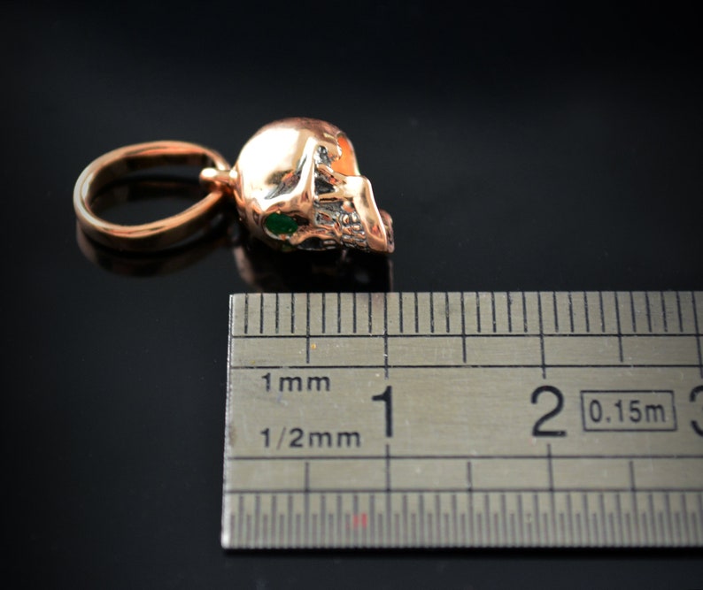 May include: A gold-colored skull pendant with green eyes, measuring approximately 1 cm in length. The pendant is shown next to a ruler for size reference.