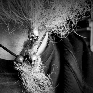 Op de afbeelding: Een zwart-witfoto van een persoon die een ketting draagt met drie zilveren schedelcharms. De ketting hangt aan een lange lok haar.