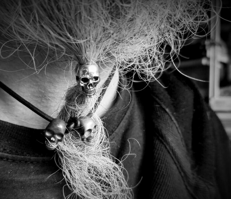 Op de afbeelding: Een zwart-witfoto van een persoon die een ketting draagt met drie zilveren schedelcharms. De ketting hangt aan een lange lok haar.