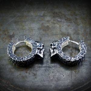 K&ouml;nnte beinhalten: Ein Paar silberne Totenkopf-Creolen mit strukturierter Oberfl&auml;che. Die Ohrringe sind aus Metall und haben einen Scharnierverschluss.