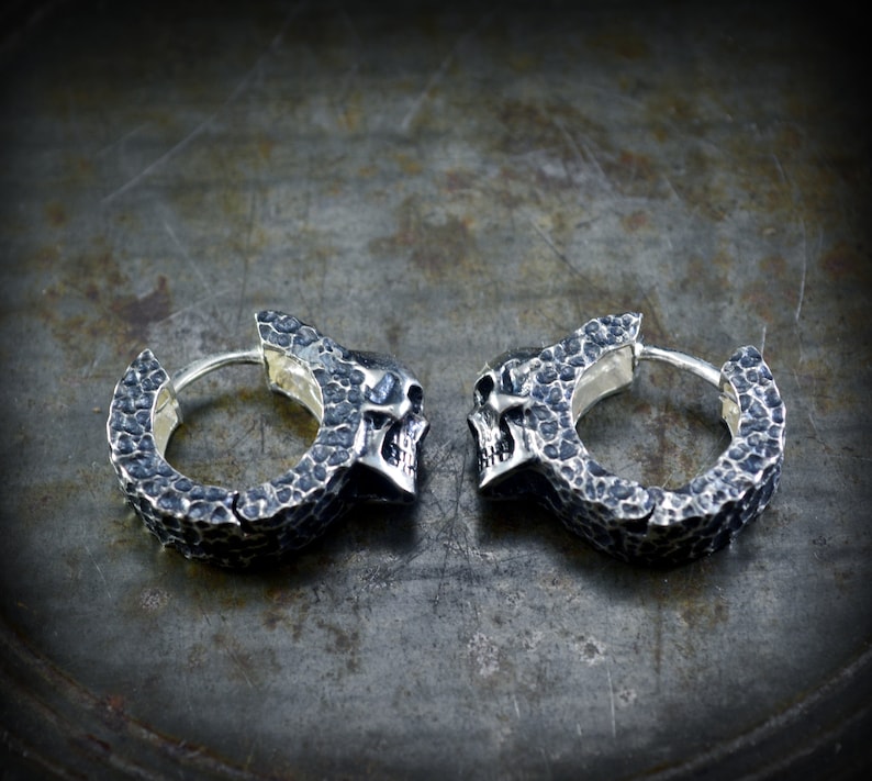 K&ouml;nnte beinhalten: Ein Paar silberne Totenkopf-Creolen mit strukturierter Oberfl&auml;che. Die Ohrringe sind aus Metall und haben einen Scharnierverschluss.