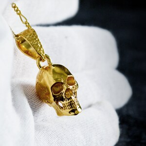 May include: A gold skull pendant necklace with a textured surface. The pendant is hanging from a gold chain.