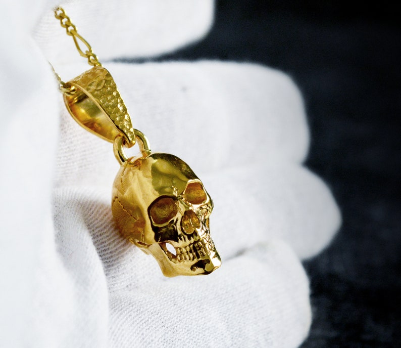 May include: A gold skull pendant necklace with a textured surface. The pendant is hanging from a gold chain.