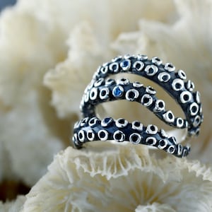 Peut inclure: Bague en argent en forme de pieuvre avec trois tentacules et des pierres précieuses bleues. La bague est posée sur un fond de corail blanc.