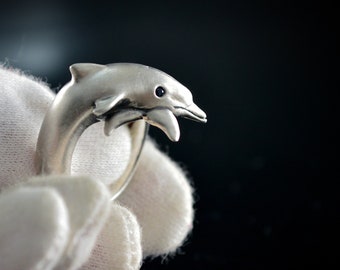 May include: A silver-toned ring featuring a dolphin design. The dolphin has a detailed head and body, with a small, dark-colored eye. The ring is held by a gloved hand against a dark background, highlighting the ring's details.