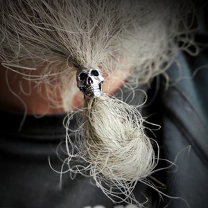 Op de afbeelding: Een zilveren schedel haarband met een gedetailleerd ontwerp. De haarband houdt een lange, witte paardenstaart vast.
