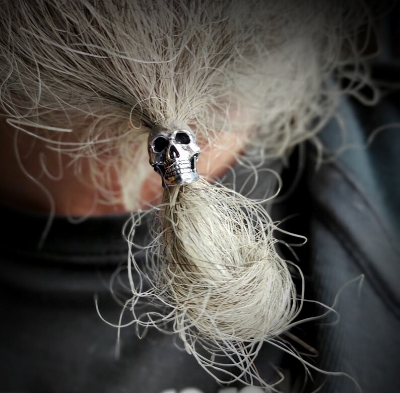 Op de afbeelding: Een zilveren schedel haarband met een gedetailleerd ontwerp. De haarband houdt een lange, witte paardenstaart vast.
