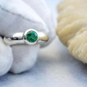 May include: A silver ring featuring a bright green gemstone. The ring has a polished band and a bezel setting. The gemstone is round. The ring is held by a gloved hand. The ring's design is simple and elegant.