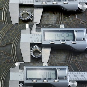 K&ouml;nnte beinhalten: Ein silberner Totenkopf-Hoop-Ohrring, der mit einer digitalen Schieblehre gemessen wird. Die Schieblehre zeigt 196,8 mm, 145,2 mm und 588 mm an.